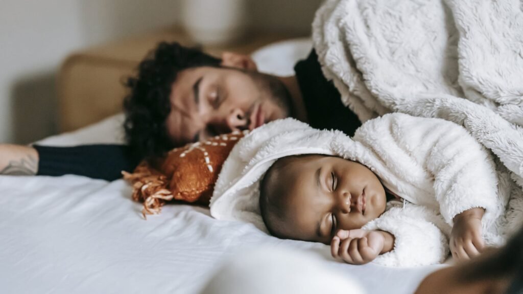 | Chill & Thrive Multiethnic father and newborn baby lying and sleeping together on comfortable bed near crop unrecognizable woman in daylight