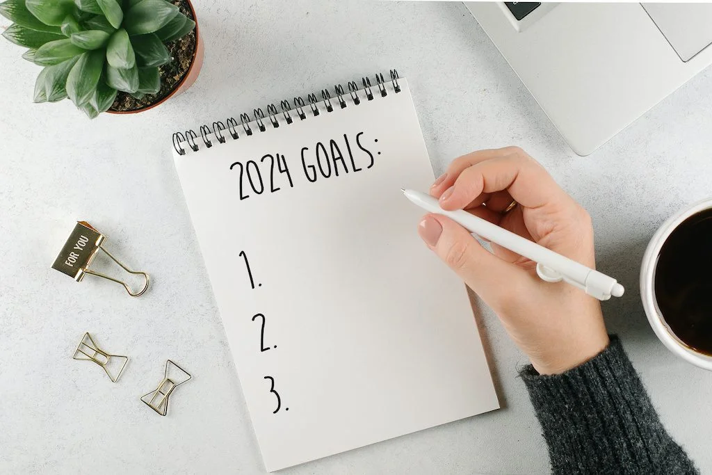 Woman's hand writing 2024 | Chill & Thrive | Chill & Thrive Womans hand writing 2024 Goals in notebook Laptop home plant cup of coffee on light table